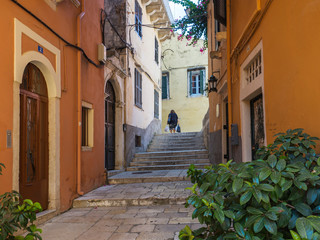 Corfu old town narrow street with older local woman carring bags with shopping purchaise, stairs, doors and windows and flower garlands, summer day, Kerkyra townCorfu island, Greece