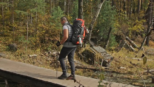 People walk on a wooden bridge over the river. Family travels. People environment by mountains, rivers, streams. Parents and kids walk using trekking poles. Man and woman have hiking backpacks, flasks