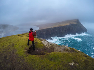 Scenic landscape view of young tourist man taking photo of high cliffs and stormy sea next to the Sorvagsvatn lake on Vágar Island. Tourist popular attraction/destination in Faroe Islands, Denmark