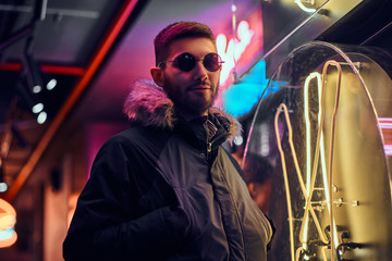 A handsome man wearing a coat and sunglasses with hands in pockets, standing in the night on the street. 