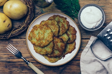 Pan-fried potato pancakes (rosti, kartoffelpuffer, latkes, draniki, hash browns): traditional potato fritters on a wooden background with sourcream and greens
