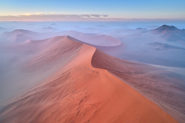 Fototapeta na wymiar Aerial, artistic photo of dunes of Namib desert covered on mist. Famous orange dunes of Namib from above. Desert landscape. Sunrise in Namib desert. Travel photography, Namib-Naukluft National Park.