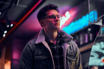 Fototapeta premium A young man fashionably dressed standing in the street at night. Illuminated signboards, neon, lights.