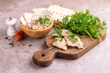 Pieces of matzah with homemade liver pate with parsley and onion on old wooden cutting board with burlap napkin. Gray marble background. Copy space
