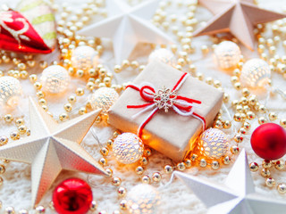 Fototapeta na wymiar Christmas and New Year star decorations on white knitted background. Present box wrapped in craft paper with snowflake symbol on it, metal light bulbs with delicate pattern, golden beads, red balls.