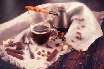 Close-up shot of cup of coffee with roasted beans, chocolate, badyan, cardamom and cezve on a rustic wooden table.