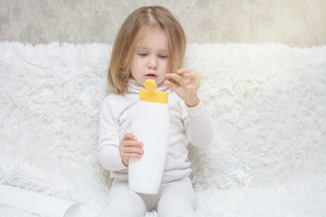 Cute little girl keeping white plastic shampoo bottle in her hands, looking at the camera. Baby bath, hygiene accessories. Cosmetic package collection for foams, shampoo © silentalex88