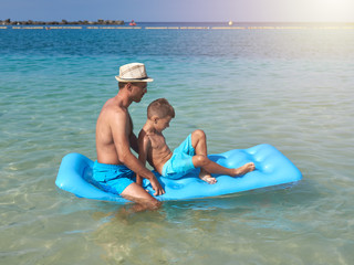 Father and son are swimming on the blue air mattress in the crystal ocean water. They are enjoying...