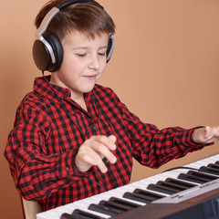 Cute European boy in a red plaid shirt is having his music classes. He is playing electric piano,...