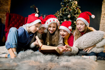 Charming and beautiful little children in Santa's hats are reading Christmas stories and laughing, on the background of a Christmas tree.