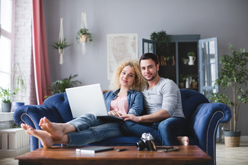 Man and woman with laptop sitting on a blue sofa at home. Young family planning.