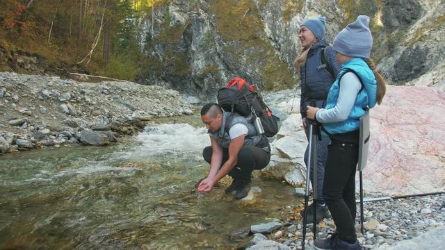 People walk near the mountain river. Family travels. People environment by mountains, rivers, streams. Parents and kids walk using trekking poles. Man and woman have hiking backpacks. Dad mom and
