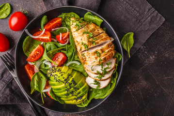 Grillwd chicken breast and avocado salad with spinach, tomatoes and Caesar dressing, dark background, top view.