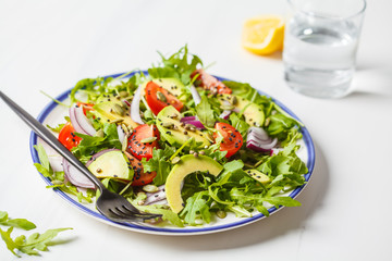 Healthy arugula, avocado, tomatoes salad on white marble background.