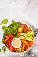 Vegan salad bowl with baked vegetables, chickpeas, avocado and tahini dressing on a white background, top view.