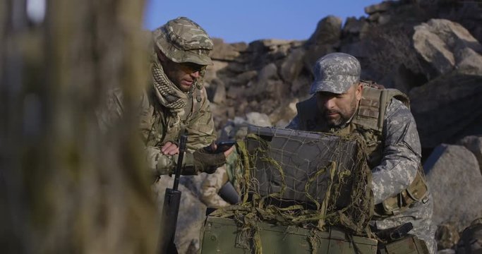 Medium shot of fully equipped and armed soldiers looking at a computer and another looking at a handheld device in the field and search coordinates