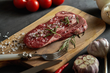 one raw piece of steak with salt, pepper on a wooden board and a set of vegetables on a black table
