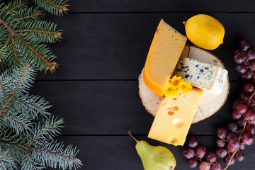 Various types of cheese and fruits on dark boards and Christmas tree branches. Swiss cheese, blue cheese, Dutch cheese for Christmas. Dairy products, lemon, grapes, pears. Top view