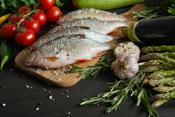 four fresh raw dorada fish on a wooden board with a set of vegetables on a black table