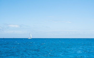 Fototapeta premium Small yacht far at the horizon with white sail, calm ocean, blue sky