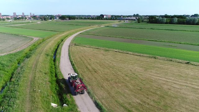 Static aerial footage of tractor driving over narrow rural road carrying retracted farm implement also showing countryside grass fields on left and right side of path beautiful summer day 4k quality