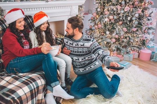 Lovely picture of family sitting together on gfloor. Girl holds bowl of popcorn. Father looks at her and uses remote control at the same time. Young woman and girl wears Christmas hats.