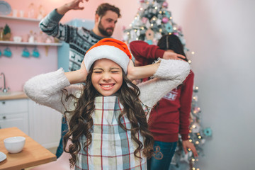 Small girl in festive clothes stands and close her ears with ahnds. She has closed her eyes. There are parents behind her. Father keeps hand up. He is going to beat his wife. He tries protect herself.