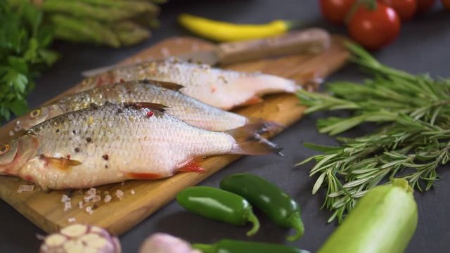 three fresh raw dorada fish on a wooden board with a set of vegetables on a black table