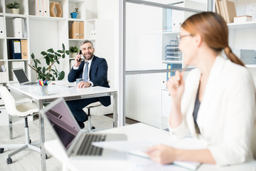 Positive confident handsome male manager in formalwear sitting at table and talking by mobile phone while conferring with colleague in office