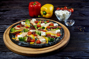 Hot aromatic tasty Italian pizza on black dough with meat, vegetables and cheese on a dark wooden board with whole paprika, a sprig of tomatoes,sliced cheese and spices.