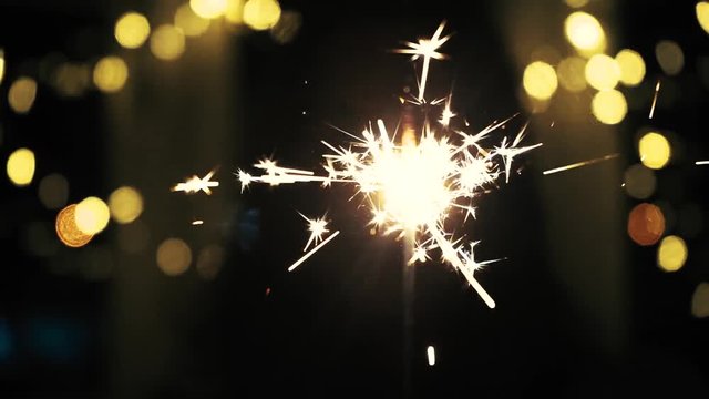 Beautiful firework sparkler burning with sparks and flames on the black background with Christmas lights. New Year concept. Close up. Slow motion