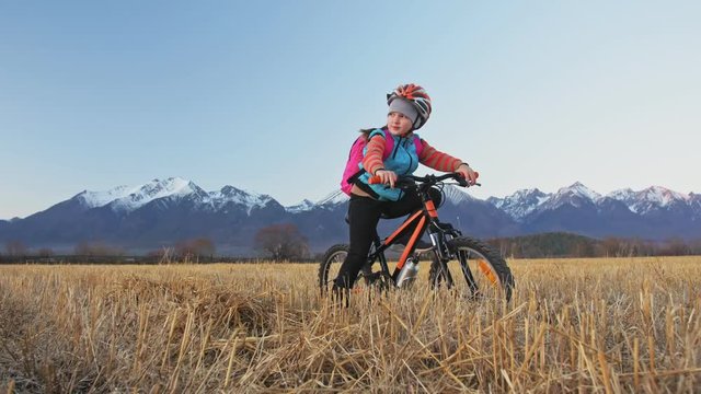 One caucasian children walk with bike in wheat field. Girl walking black orange cycle on background of beautiful snowy mountains. Biker motion ride with backpack and helmet. Mountain bike hardtail.