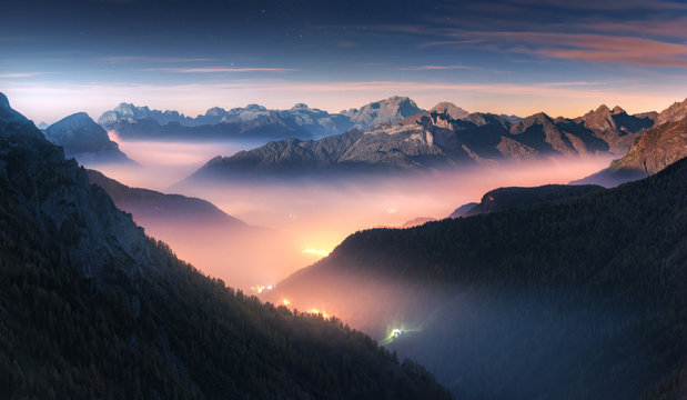 Mountains in fog at beautiful night in autumn in Dolomites, Italy. Landscape with alpine mountain valley, low clouds, forest, colorful sky with stars, city illumination at dusk. Aerial. Passo Giau