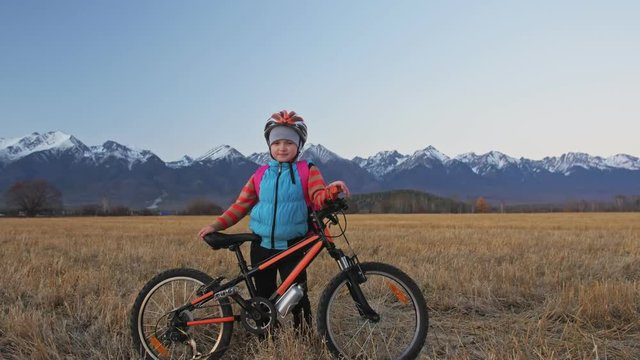 One caucasian children walk with bike in wheat field. Girl walking black orange cycle on background of beautiful snowy mountains. Biker motion ride with backpack and helmet. Mountain bike hardtail.