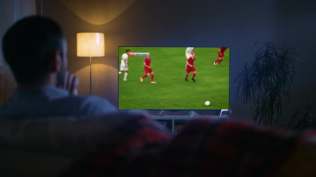 At Home In the Living Room Soccer Fan Relaxes on a Sofa, Watches TV, His Favourite Soccer Team Playing Professionally, Scoring a Goal. 