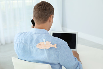 Man with paper fish on his back working with laptop at table indoors. April fool's day prank