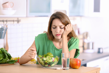 Unhappy woman eating vegetable salad at table in kitchen. Healthy diet
