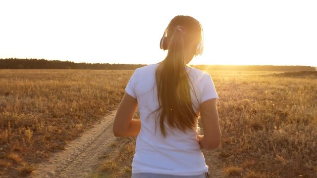 Beautiful girl doing sports exercises at sunset in countryside. girl in headphones runs along road and listens to music. view from back