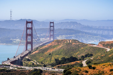 Aerial view of Golden Gate Bridge and the freeway bordered by the green hills of Marin Headlands on a sunny morning; San Francisco covered in a light fog layer in the background; California