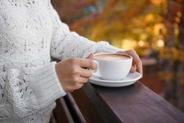Woman in warm sweater holding cup with hot cozy drink on wooden railing at balcony