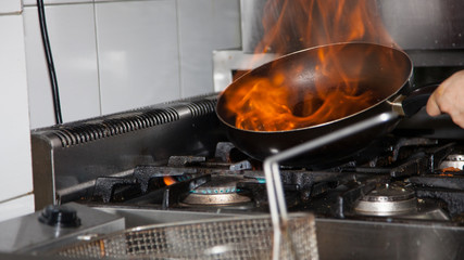 Chef Cooking With Fire In Frying Pan. Professional chef in a commercial kitchen cooking flambe style. Chef frying food in flaming pan on gas hob in commercial kitchen