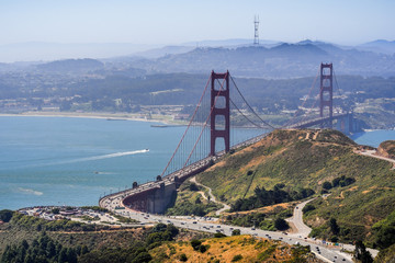 Aerial view of Golden Gate Bridge and the freeway bordered by the green hills of Marin Headlands on a sunny morning; San Francisco covered in a light fog layer in the background; California
