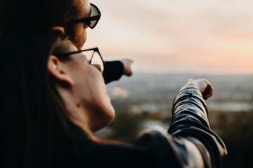 Crop man and woman in glasses pointing at distance together while standing on blurred background of nature during sunset. Young couple pointing at distance