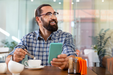 Some sugar. Bearded smiling man putting some sugar into his coffee sitting in cafeteria holding his phone