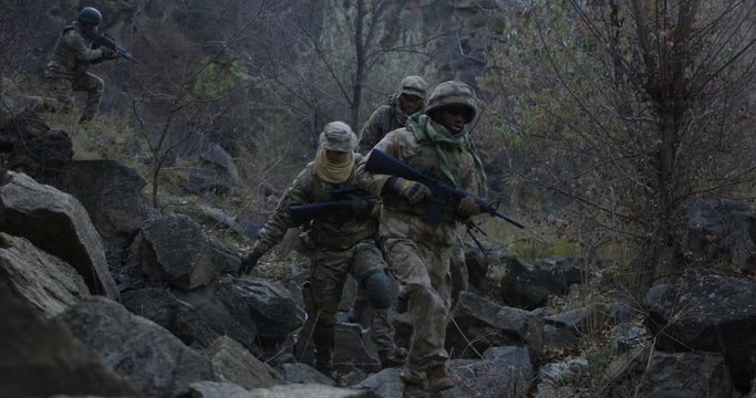 Medium slow motion shot of fully equipped and armed soldiers walking in single file through rocks at dusk