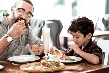 Beaming son. Funny beaming curly dark-haired son feeling happy eating cheesy pizza with father