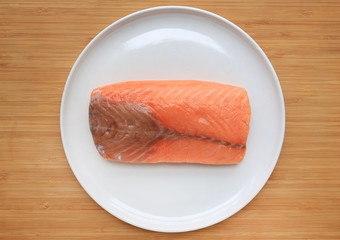 Raw piece of sliced salmon on white plate against wooden board background.