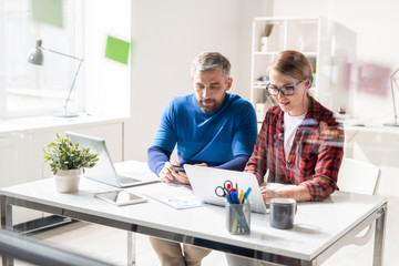 Serious busy team of app developers in casual clothing sitting at table with office supplies and using modern devices while testing new app