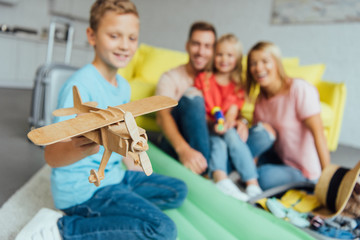 boy playing with toy wooden airplane with family having fun on background and packing for summer holiday
