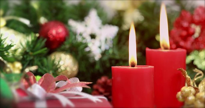 Close up scene VDO of red candles were lit, gift box on foreground, pine branches with beautiful ornament behind it, Christmas theme.